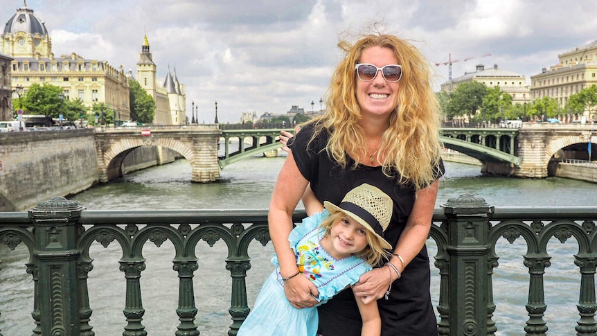 A woman wearing sunglasses hugs a child wearing a hat on a bridge.