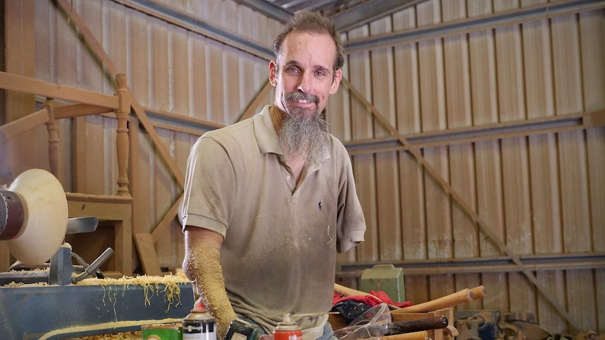 Dale Cordwell in his wood turning shed, he is sitting on a table with a wood lathe machine behind him.