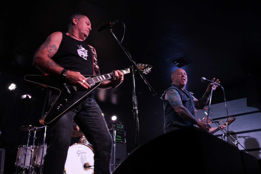 Two men on stage, one with a guitar, one bald, both with tattoos