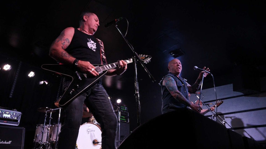 Two men on stage, one with a guitar, one bald, both with tattoos