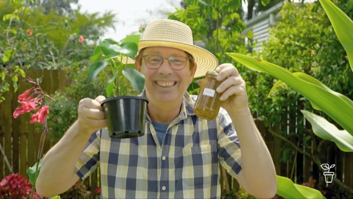 Man in hat smiling and holding up a pot plant in one hand and a jar of homemade jam in the other