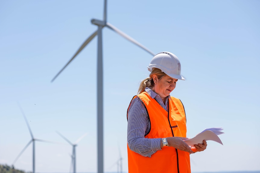 The premier in a hard hat and fluro vest looking down at papers with a wind turbine in the background.