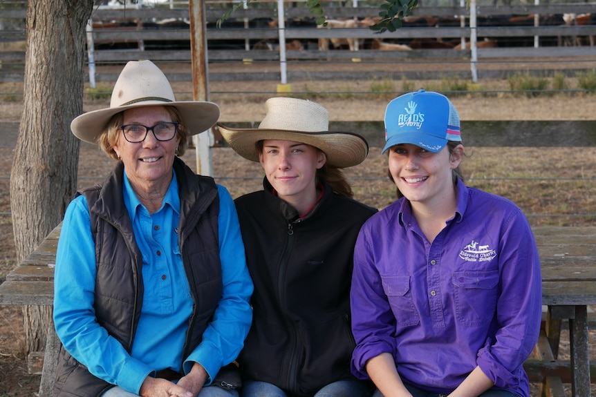 A woman in a blue work shirt and hat sits to the left of two girls wearing hats, all smiling.