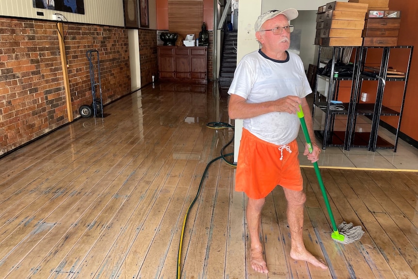 A man with grey hair and moustache mops the wooden floor while wearing orange pants and a white hat. 