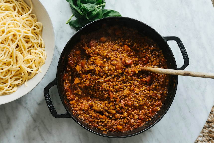 Cast iron pot containing lentil bolognaise ragu, with a bowl of spaghetti and fresh basil beside it.