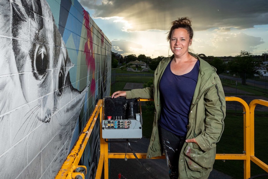 A smiling woman on scissor lift beside a brick wall featuring a large painted sugar-glider possum as the sun sets behind