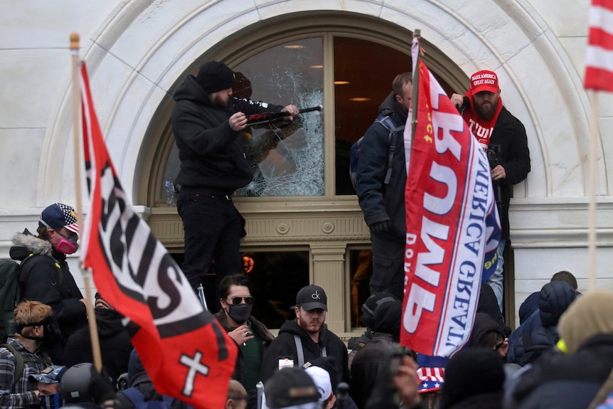 Young men, wearing black and bearing red Trump flags and banners, gather before a doorway at the Capitol, one smashing a window.