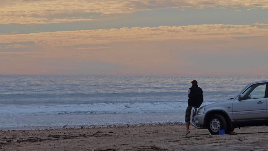 A man sits on the bonnet of a car watching the sunset on Aldinga Beach.