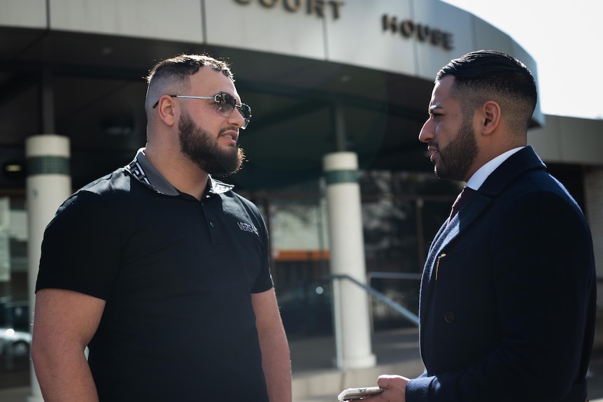 Two men talk to each other in front of a court house. One is wearing sunglasses, the other a suit and tie.