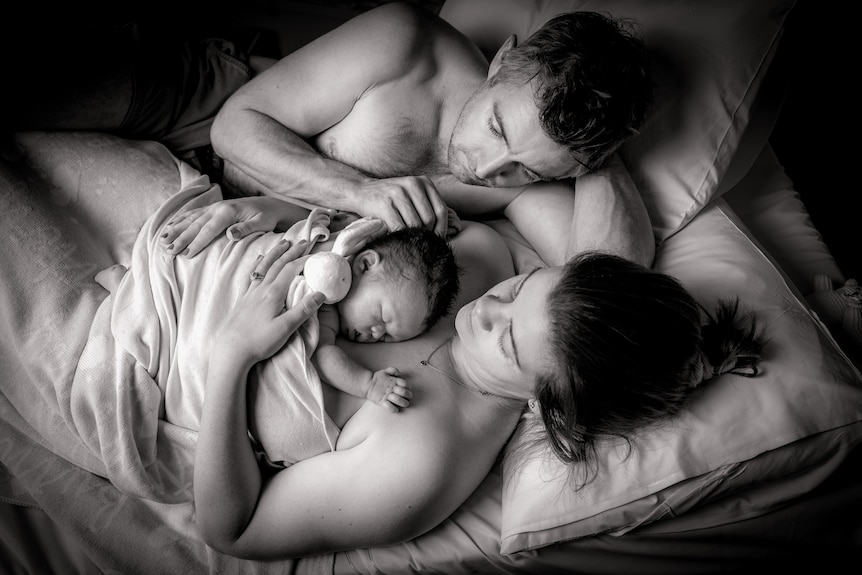 A black and white image showing parents laying on a bed, with the mother cradling a newborn baby on her chest.