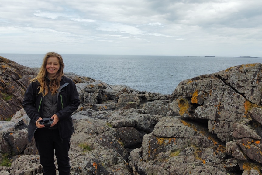 Melanie Finch hält ihre Kamera mit Felsen und Meer im Hintergrund