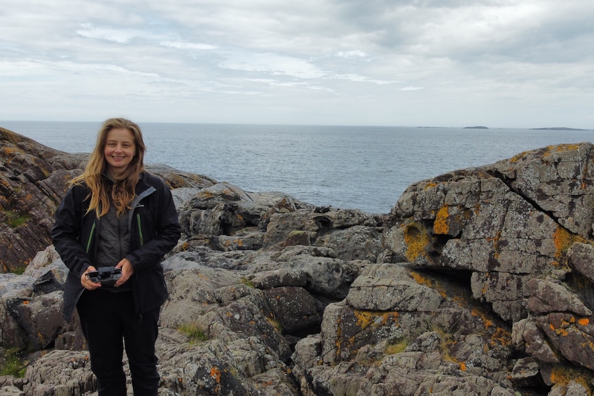 Melanie Finch holds her camera with rocks and ocean in the background