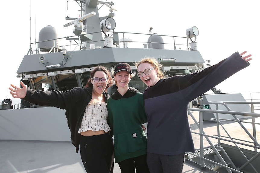 Three teenage girls on board a naval ship stand together and smile.