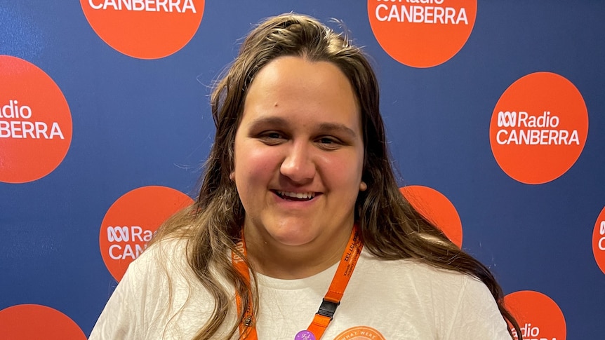 Sarah smiling at the camera in white shirt and in front of an ABC Radio Canberra red and blue banner.