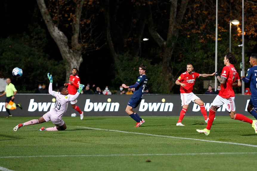 The ball flies past a diving goalkeeper while footballers watch on