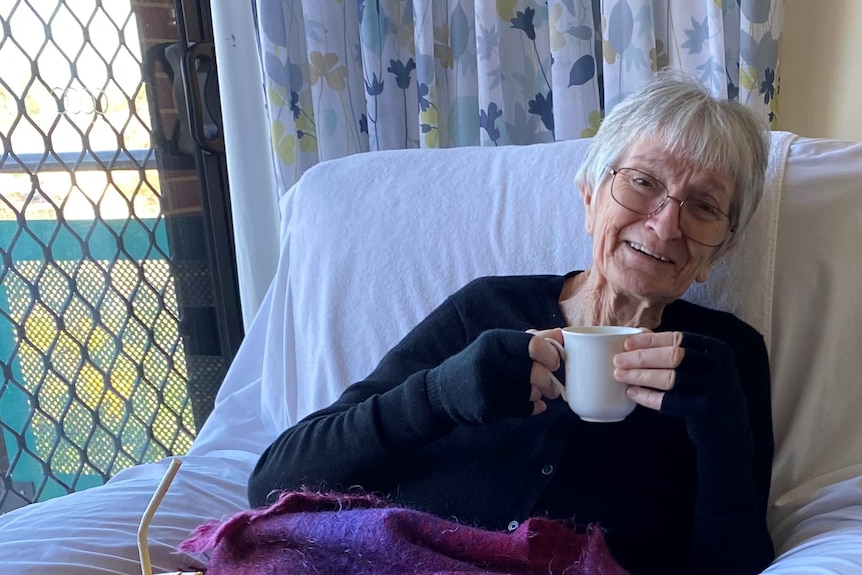 An elderly woman holds a cup of tea and smiles.