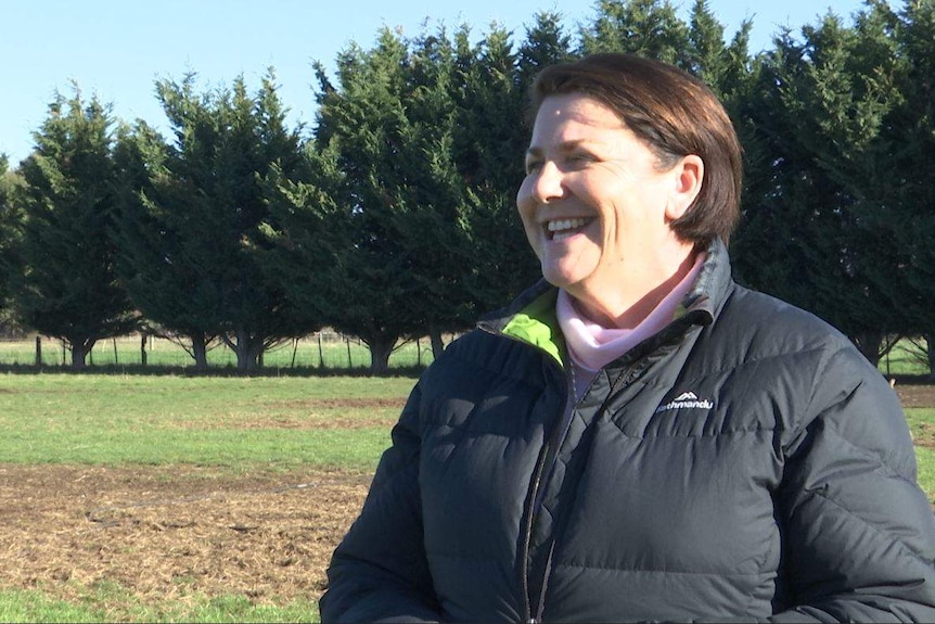A smiling woman in a puffa jacket in a field lined with pine trees.