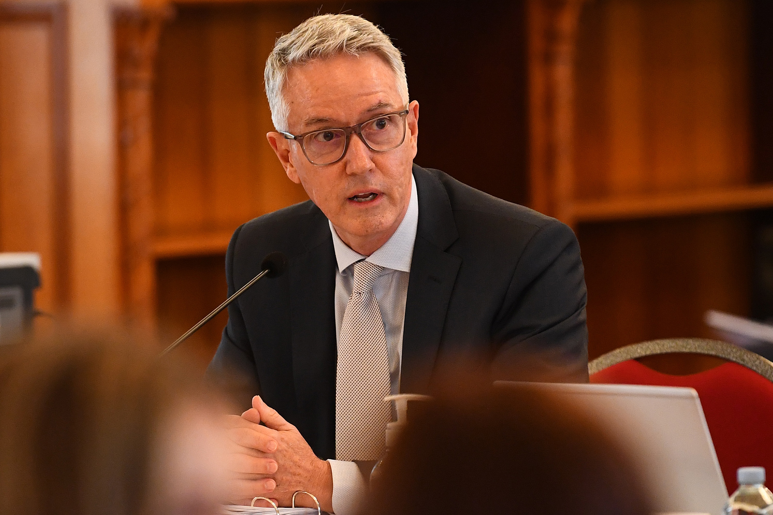 Un hombre con traje y gafas habla por un micrófono en una audiencia