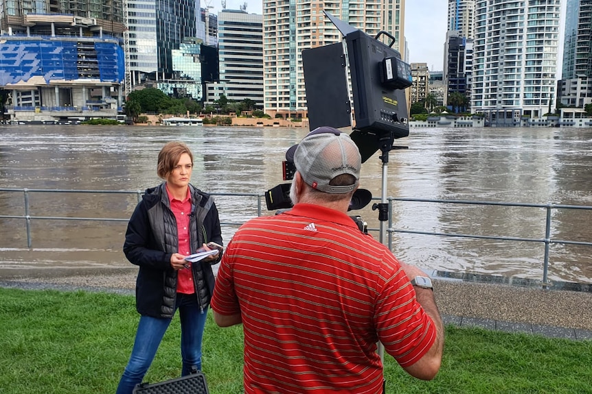 Woman standing in front of camera on a tripod with cameraman operating. River in background.