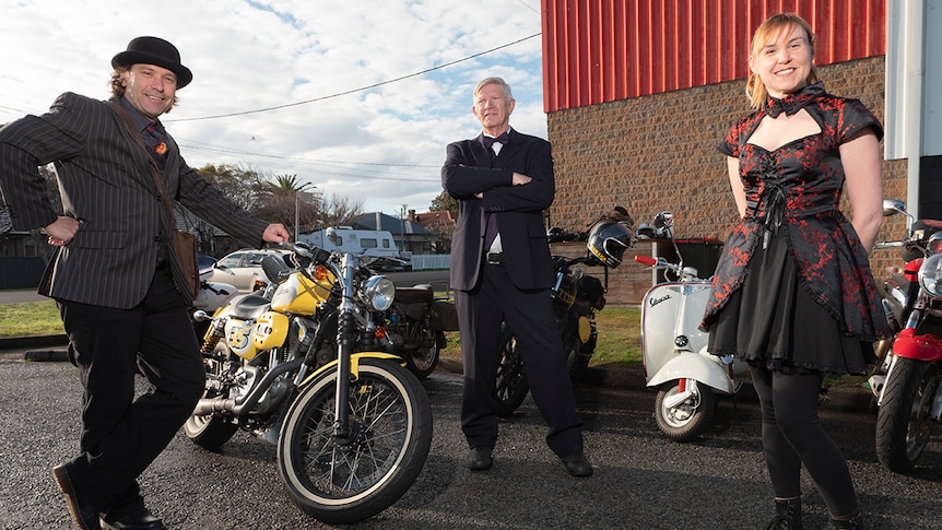 Two men and a woman dressed in fancy cloths pose in front of motorcycles outside a cafe