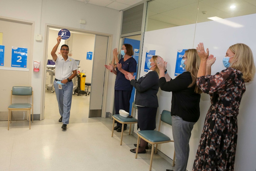 Man walks out of a surgery door holding a sign showing his is the first person to receive the covid-19 vaccine.