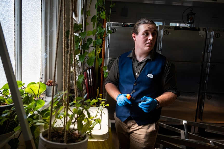 A man stands with a lighter about to light a candle. He is surrounded by plants and a mortuary freezer is in background