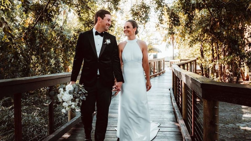 A smiling couple in wedding outfits look at each other as they walk across a wooden bridge.