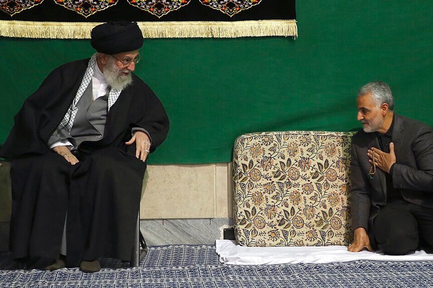 Ayatollah Ali Khamenei sits in black robes as Qassem Soleimani kneels down before him in front of a green backdrop.