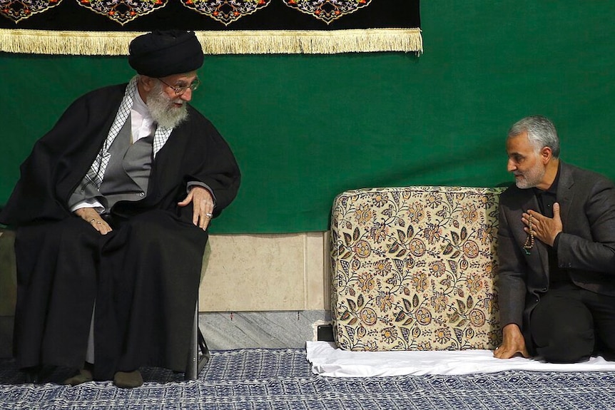Ayatollah Ali Khamenei sits in black robes as Qassem Soleimani kneels down before him in front of a green backdrop.