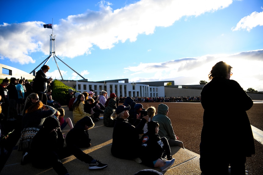 people sit together on a step in the day outside parliament house when the flag in the distance