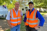 Two men standing in a caravan park wearing his vis orange vests looking at the camera