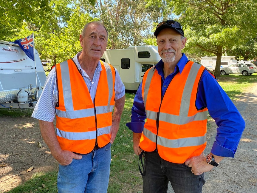 Two men standing in a caravan park wearing his vis orange vests looking at the camera