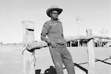 An Aboriginal stockman in the Kimberley leans on a wooden fence. Circa 1950s