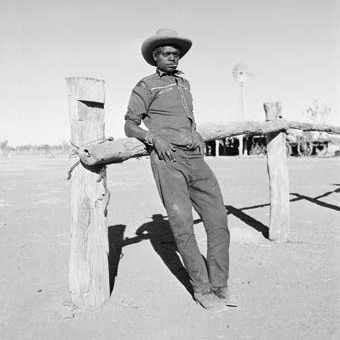 An Aboriginal stockman in the Kimberley leans on a wooden fence. Circa 1950s