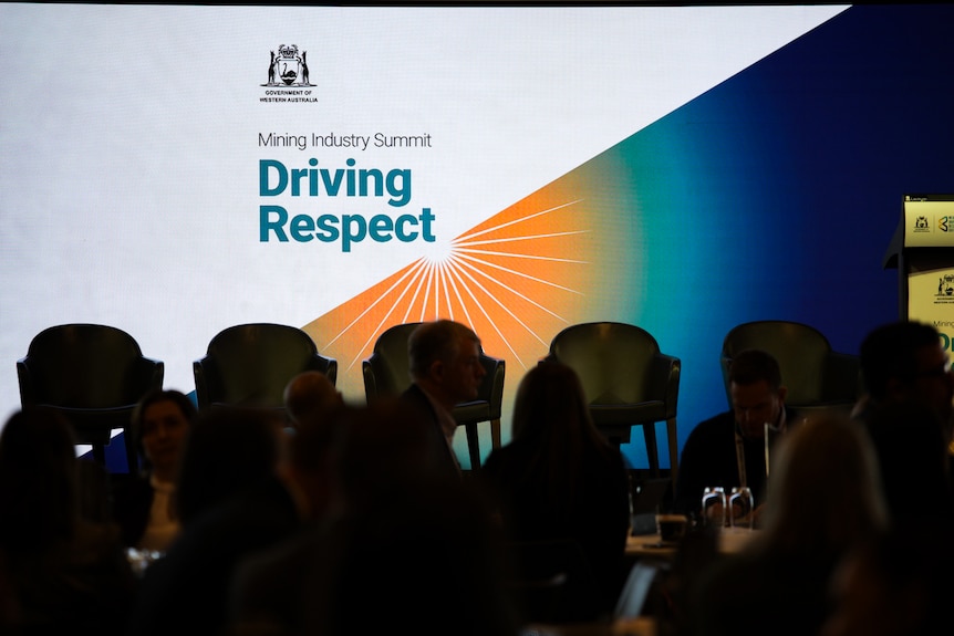 A sign that says 'Mining Industry Summit Driving Respect' with people sitting in the foreground.