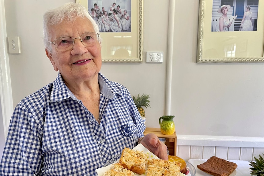 A lady with grey hair holds up a plate of slice with coconut on it.