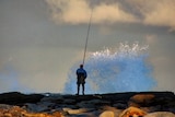 Fisherman on the rocks as a wave crashes nearby.