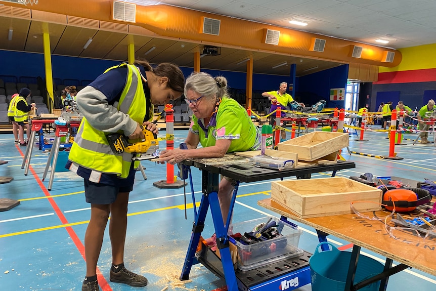 A girl in a high vis vest stands holding a jigsaw, guided by a woman in a fluoro shirt.