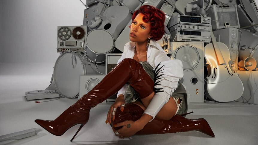 Raye sitting down wearing long maroon high heeled boots, a grey guitar, drum kit and speakers behind her