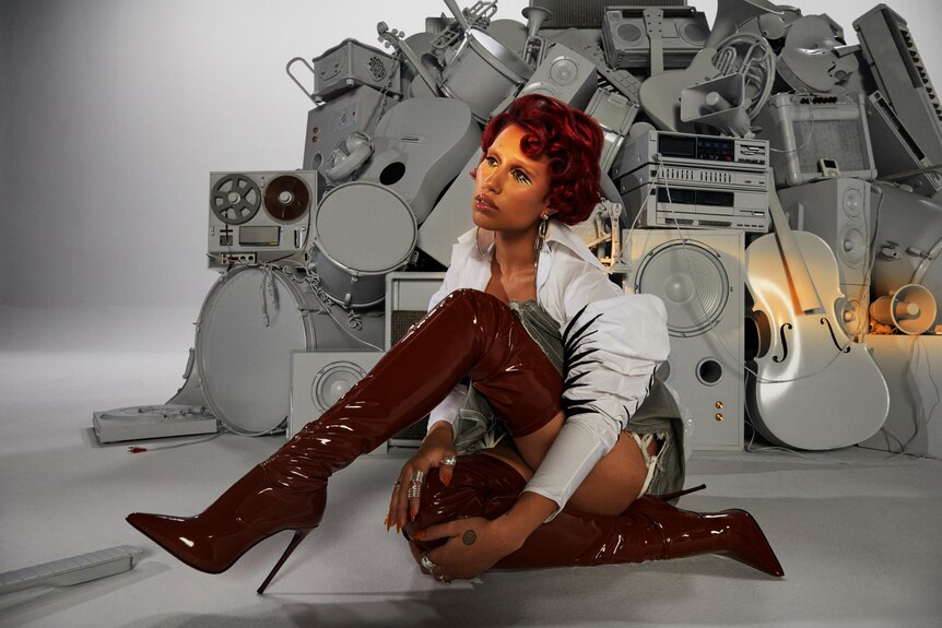 Raye sitting down wearing long maroon high heeled boots, a grey guitar, drum kit and speakers behind her