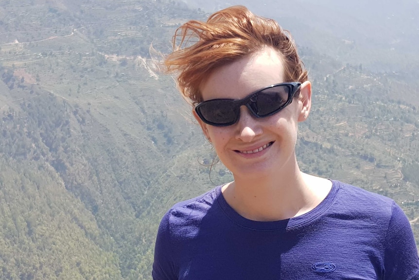 A woman with short red hair and sunglasses and a purple shirt with a view from a mountain in the background.