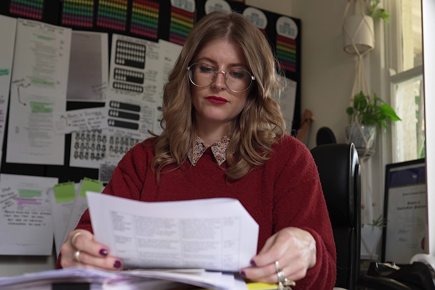 A young woman in a red jumper and large round glasses sits at a desk with lots of papers on it