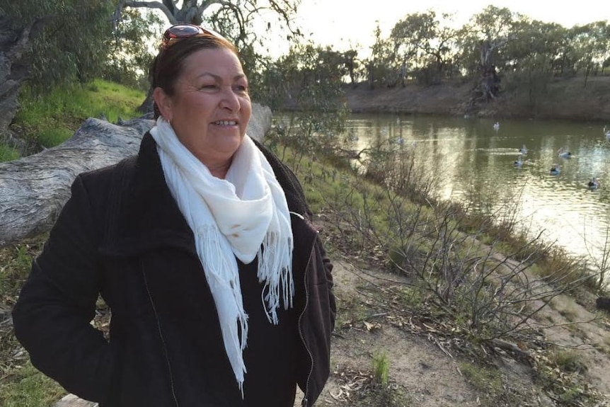 A woman wearing a black coat and white scarf stands beside the Murray River surrounded by trees.