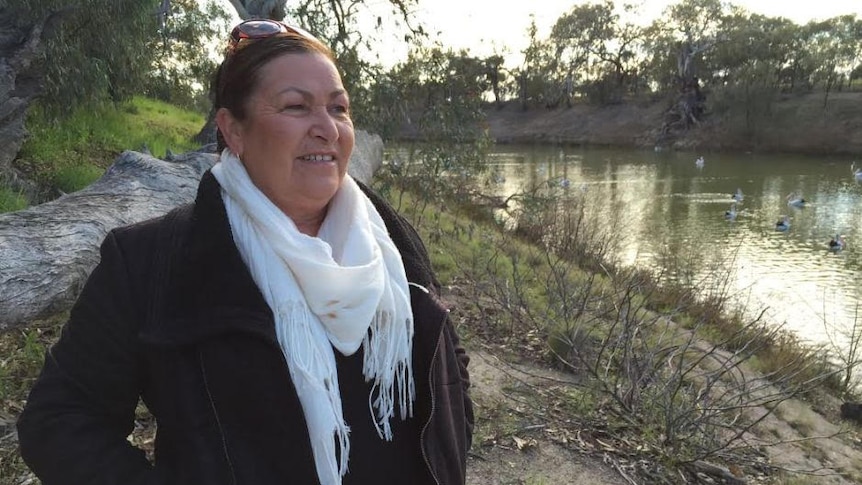 A woman wearing a black coat and white scarf stands beside the Murray River surrounded by trees.
