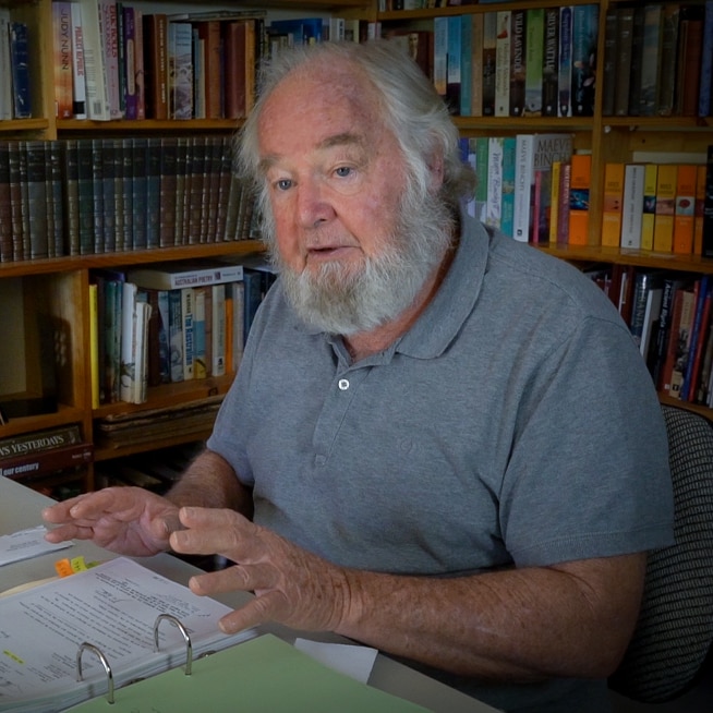 An older man with beard in his home study leafing through a folder of papers.