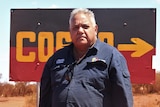 He stands in a dark blue shirt in front of a sign saying 'Cosmo' painted like the Aboriginal flag
