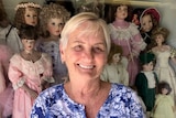 Woman with short blonde hair smiling in front of a glass cabinet of porcelain dolls