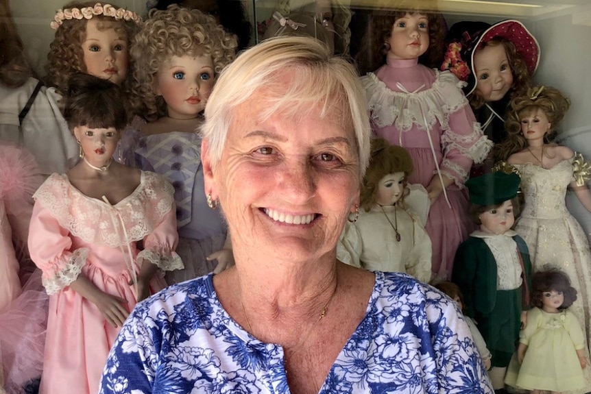 Woman with short blonde hair smiling in front of a glass cabinet of porcelain dolls