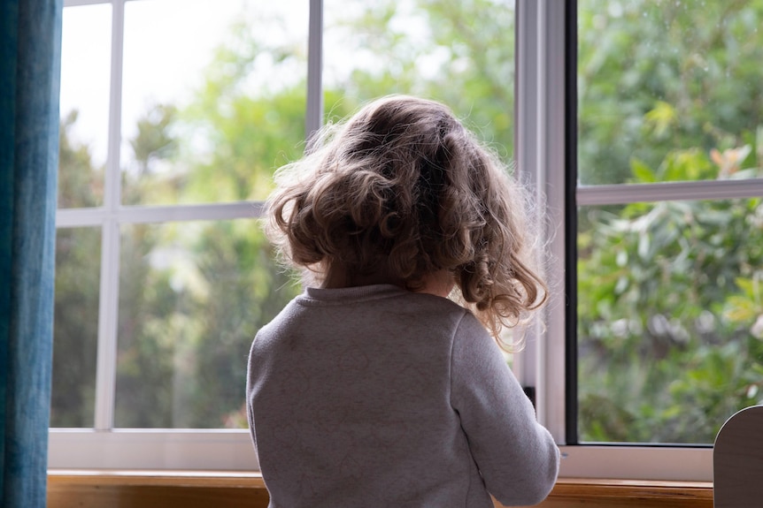 A small child stands before a bedroom window.