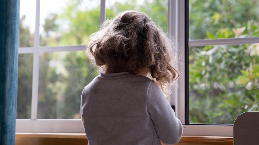 A small child stands before a bedroom window.